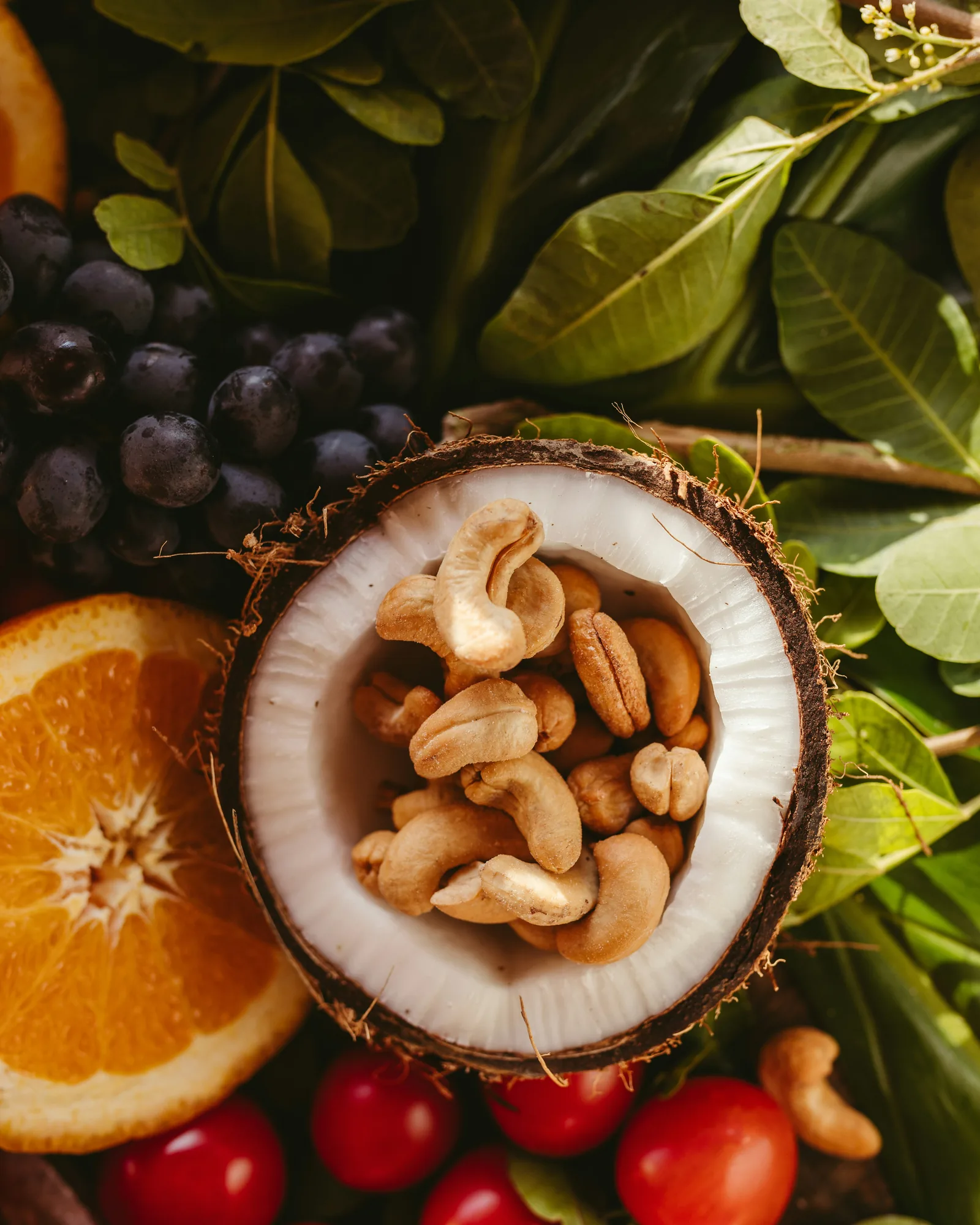 Halbe Kokosnuss voller Cashews zwischen Orangen, Trauben, Kirschtomaten und grünen Blättern, von oben